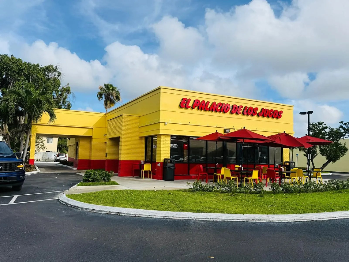 El Palacio de los Jugos location with yellow walls and red base. It features an outdoor seating area with red umbrellas and a drive-thru lane.
